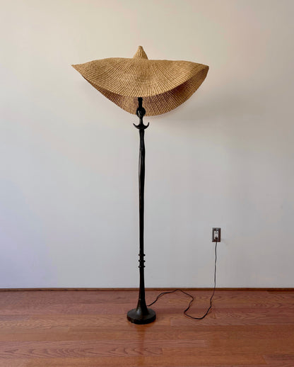 A vintage, iron 'Tête de femme' style floor lamp in the manner of Alberto Giacometti, paired with a Twenty One Tonnes handwoven, wide brimmed, natural colored light shade.