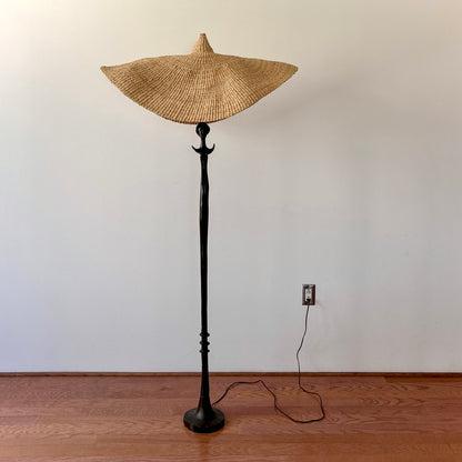 A vintage, iron 'Tête de femme' style floor lamp in the manner of Alberto Giacometti, paired with a Twenty One Tonnes handwoven, wide brimmed, natural colored light shade.