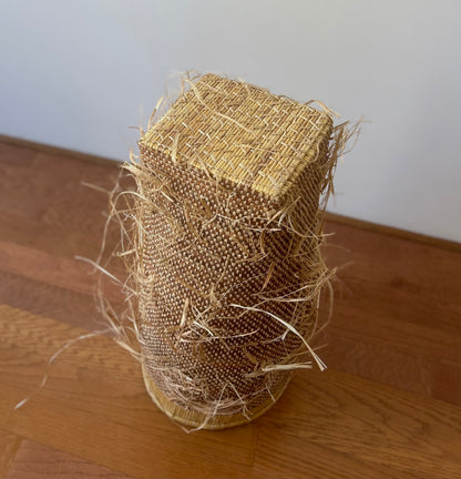 Showing the bottom of a tall storage basket, natural colored with a shaggy textured exterior. Made in Ghana from local sustainable materials.