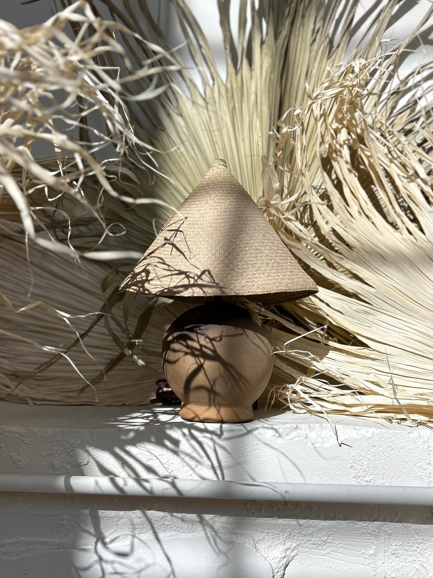 The Ash Lamp has a handcrafted, round, terracotta colored ceramic base and a cone shaped, hand woven, dried palm leaf lampshade - available in either a natural green or blonde colour. 