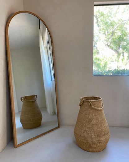 A mirror and an oversized pear-shaped handwoven basket, with two handles. Made in Ghana from local sustainable grass.