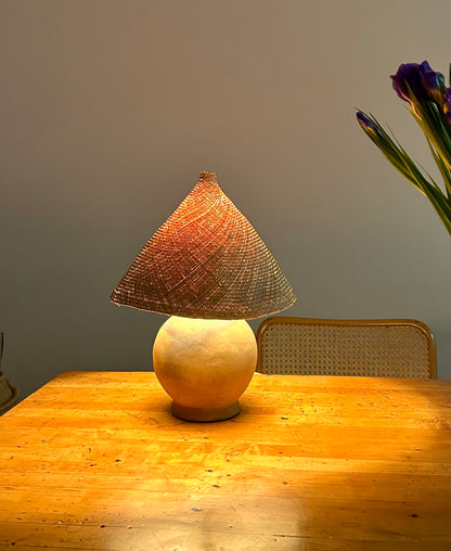 The Ash Lamp has a handcrafted, round, terracotta colored ceramic base and a cone shaped, hand woven, dried palm leaf lampshade - available in either a natural green or blonde colour. Shown here lit up on a table.