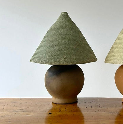 The Ash Lamp has a handcrafted, round, terracotta colored ceramic base and a cone shaped, hand woven, dried palm leaf lampshade - available in either a natural green or blonde colour. Here the lamp is hown with a natural green shade.