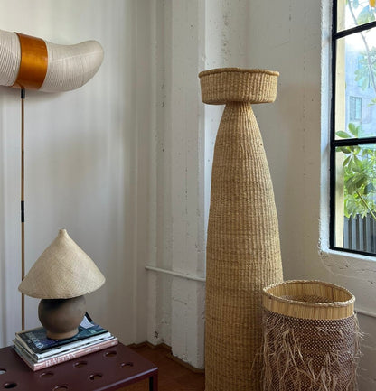 The Ash Lamp has a handcrafted, round, terracotta colored ceramic base and a cone shaped, hand woven, dried palm leaf lampshade - available in either a natural green or blonde colour. Shown here with other woven Twenty One Tonnes products.