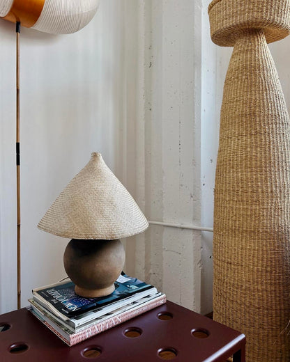 The Ash Lamp has a handcrafted, round, terracotta colored ceramic base and a cone shaped, hand woven, dried palm leaf lampshade. This shade is shown in the natural blonde colour. 