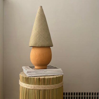 The Duende Lamp has a handcrafted, round, terracotta colored, ceramic base, with a natural colored, pointed cone shaped, hand woven, dried palm leaf lampshade. 