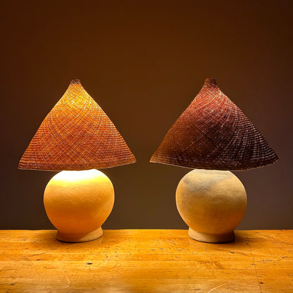 The Ash Lamp has a handcrafted, round, terracotta colored ceramic base and a cone shaped, hand woven, dried palm leaf lampshade - available in either a natural green or blonde colour. Two of them are shown here lit up.