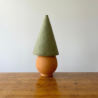 The Duende Lamp has a handcrafted, round, terracotta colored, ceramic base, with a natural colored, pointed cone shaped, hand woven, dried palm leaf lampshade. Shown here with a green colored shade.