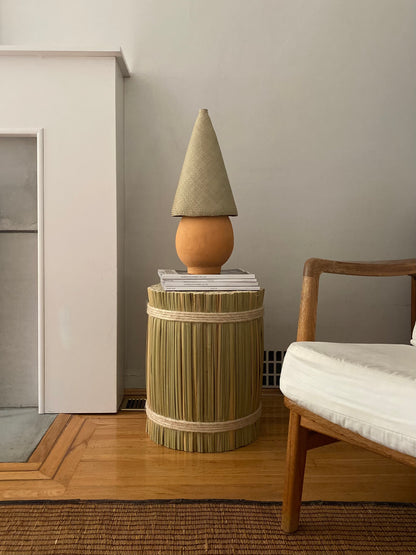 The Duende Lamp has a handcrafted, round, terracotta colored, ceramic base, with a natural colored, pointed cone shaped, hand woven, dried palm leaf lampshade. Shown here on a table.