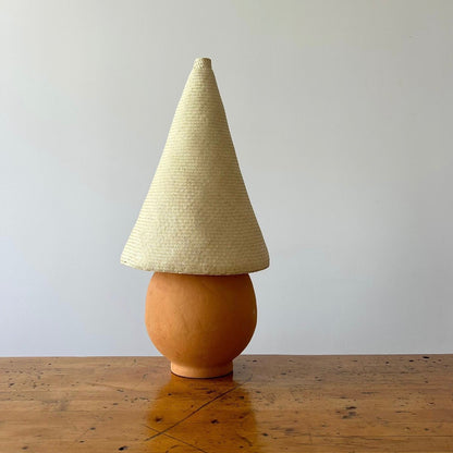 The Duende Lamp has a handcrafted, round, terracotta colored, ceramic base, with a natural colored, pointed cone shaped, hand woven, dried palm leaf lampshade. Shown here with a blonde colored shade.