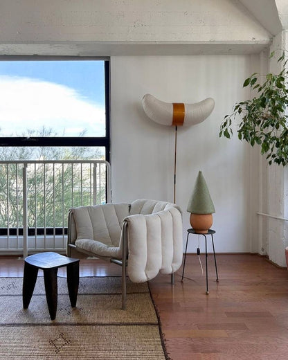 The Duende Lamp has a handcrafted, round, terracotta colored, ceramic base, with a natural colored, pointed cone shaped, hand woven, dried palm leaf lampshade. Shown here with a green colored shade in a living room.