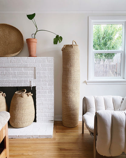 Very tall column-shaped basket with two handles, shown in a living room.  Hand crafted in Ghana from sustainable local grass.