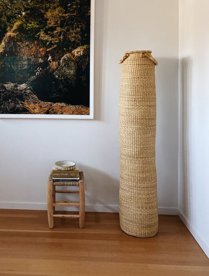 Very tall column-shaped baskets with two handles. More sculpture than storage, shown next to a stool.